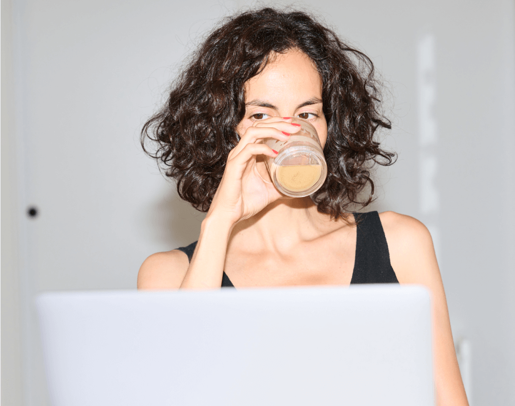 Young Woman with Dark Curly Hair Taking a Sip of Her Coffee Looking at her Laptop